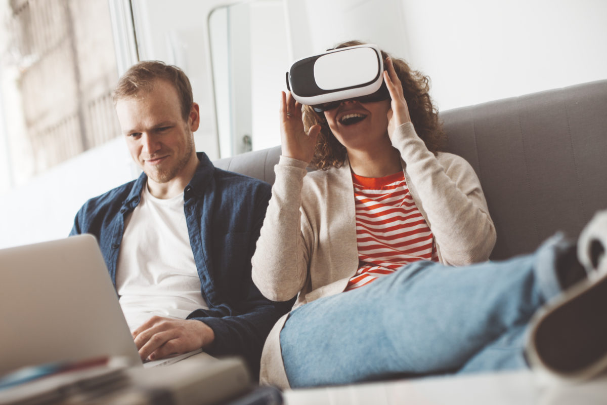 Young couple using electronic devices. Woman watching breathtaking computer game with VR headset. Man typing on laptop. Happy family life