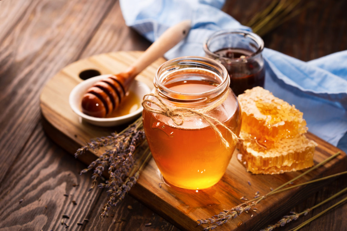 Jar of liquid honey with honeycomb inside and bunch of dry lavender over old wooden table. Dark rustic style, selective focus