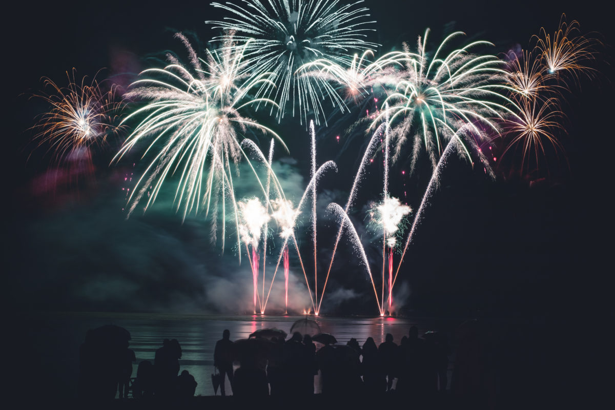 Color photography at night of beautiful large and colorful fireworks display with awe pyrotechnic effects at edge of water with some abstract long exposure reflections in water wave and with unrecognizable crowd view from back, rear view of people silhouette watching the show, during celebration event like new year or national day. This picture was taken during the Bastille Day celebrations in France on 14th of July, along Rhone river in Isere department in Auvergne-Rhone-Alpes region in Europe in Montalieu-Vercieu city.