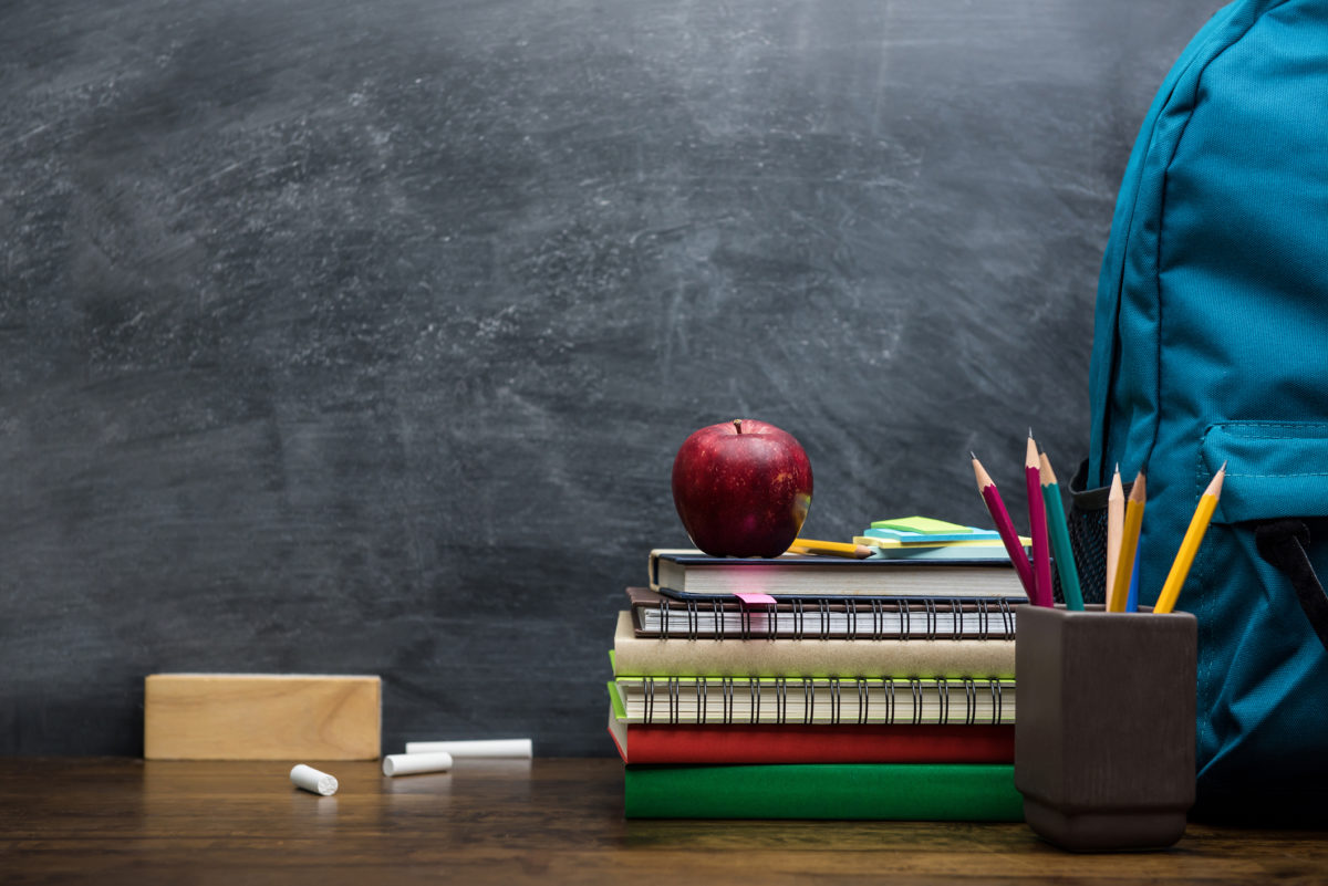 Stack of books, colorful stationery and education supplies on wooden table in classroom with blackboard in background