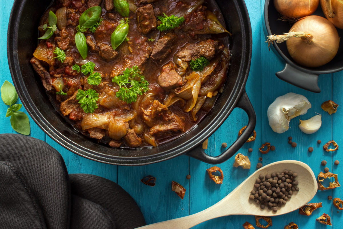 Oven cooked meat casserole in cast iron dutch oven with herbs on turquoise wooden background , top view